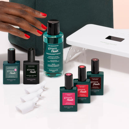A hand with red nail polish reaches for the Manucurist Starter Kit, which includes a gel polish kit, remover, clips, and a white LED lamp on a clean white surface.