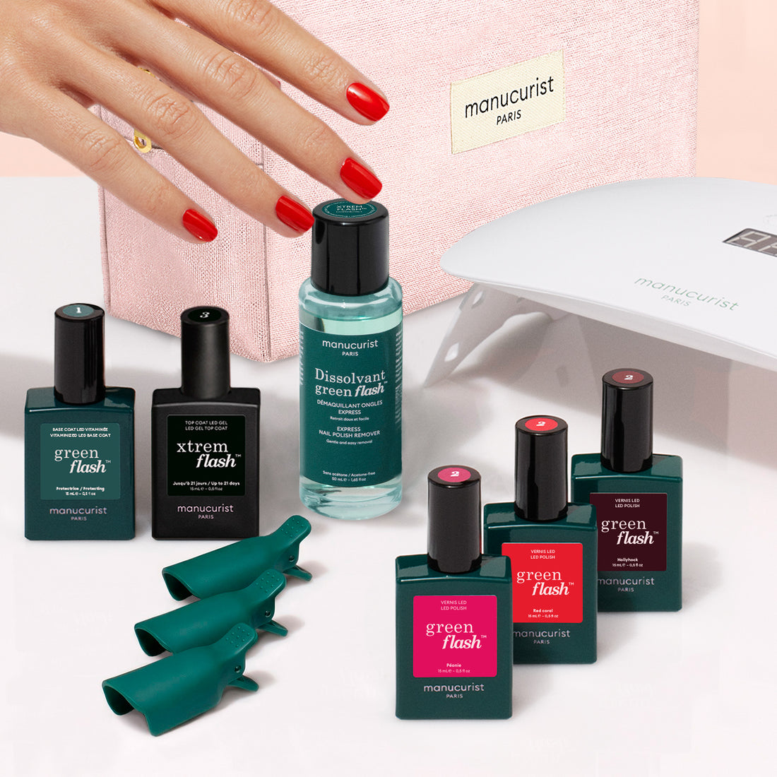 A hand with red nails touches a pink pouch among Manucurist Starter Kit items—including polish bottles, remover, clips, and a UV lamp—with the Green Flash gel polish and vitaminized base coat elegantly arranged on a white surface as part of this chic gel polish kit.