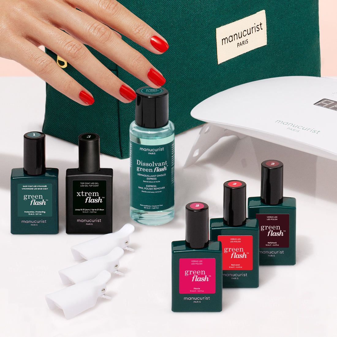 A manicured hand with red nails appears beside the Manucurist Starter Kit, featuring nail polish bottles, remover, nail clips, a UV lamp, and a green pouch—neatly arranged on a light surface for an ultra-glossy manicure.