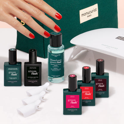 A manicured hand with red nails appears beside the Manucurist Starter Kit, featuring nail polish bottles, remover, nail clips, a UV lamp, and a green pouch—neatly arranged on a light surface for an ultra-glossy manicure.