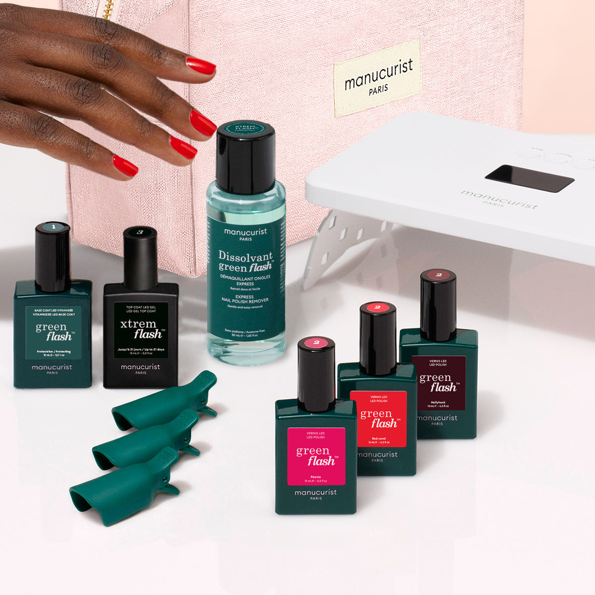 A hand with red nails reaches toward the Manucurist Starter Kit, featuring nail polish, remover, green finger covers, a white LED nail lamp, and a sleek vitaminized base coat—all arranged on a white surface next to a pink case.