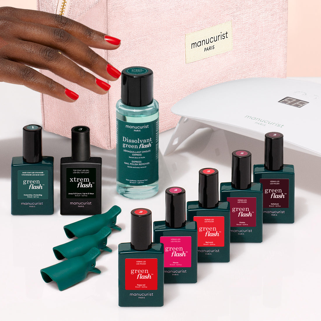 A hand with red nails reaches towards the Manucurist Super Starter Kit, featuring Green Flash gel polish, remover, finger caps, a vitaminized base coat, a nail lamp, and a pink bag labeled manucurist PARIS—perfect for achieving 21 days’ wear.