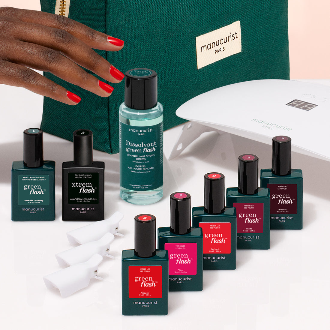 A hand with red nails reaches for the Manucurist Super Starter Kit, featuring Green Flash gel polish, Vitaminized Base Coat, white nail clips, a manicure lamp, and a green pouch—everything needed for an ultra-glossy manicure.