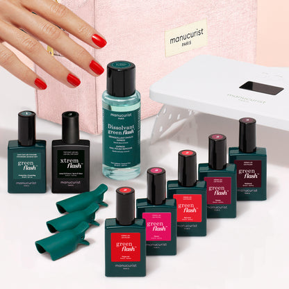 A hand with red nails reaches toward the Manucurist Super Starter Kit, which features Green Flash gel polish bottles, a vitaminized base coat, nail clips, and a white manicure lamp—all displayed on a pink surface with a pink box in the background—promising up to 21 days of flawless wear for your manicure.