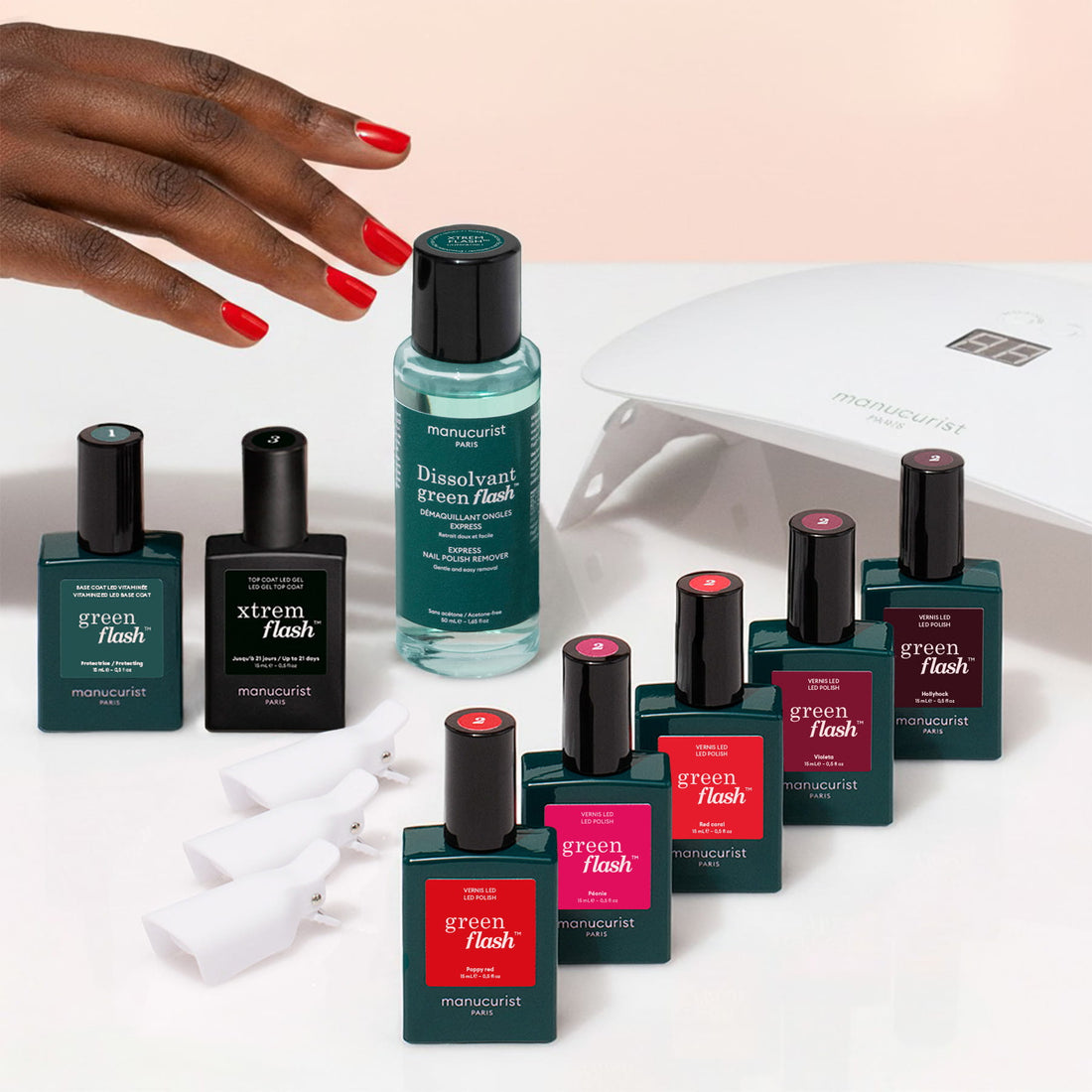A hand with red nails reaches toward Manucurist’s Super Starter Kit, featuring a gel polish kit, nail polishes, remover bottle, nail clips, and an LED lamp—all arranged on a light surface—for the perfect ultra-glossy manicure.