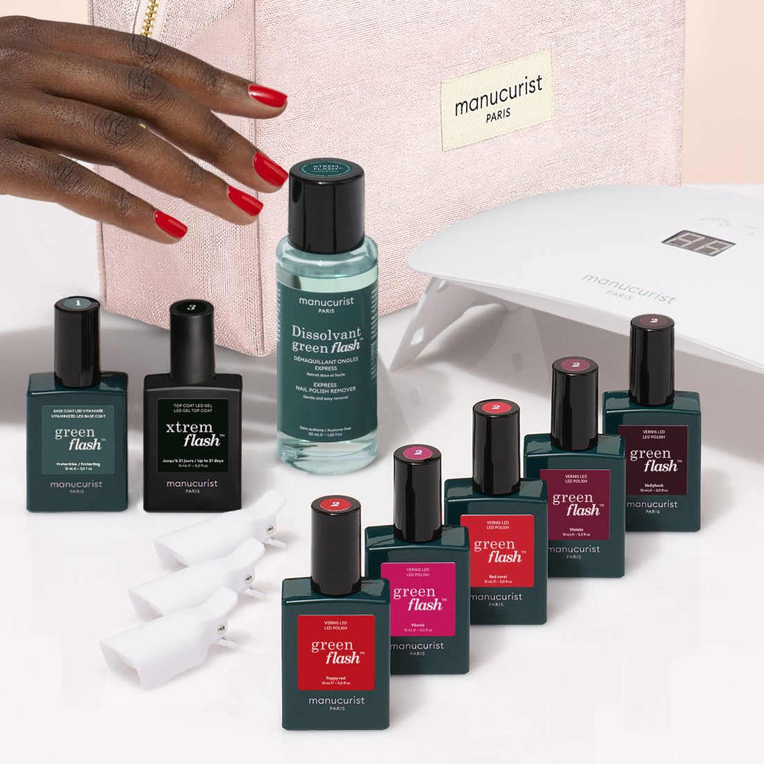 A hand with red-painted nails reaches toward the Manucurist Super Starter Kit, featuring Green Flash gel polish, remover, manicure tools, a pink pouch, and an LED lamp for an ultra-glossy manicure.