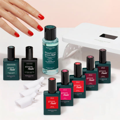 A hand with red nails, several bottles from the Manucurist Super Starter Kit, a remover, nail clips, and a nail lamp are arranged on a light surface—everything you need from Manucurist for an ultra-glossy manicure.