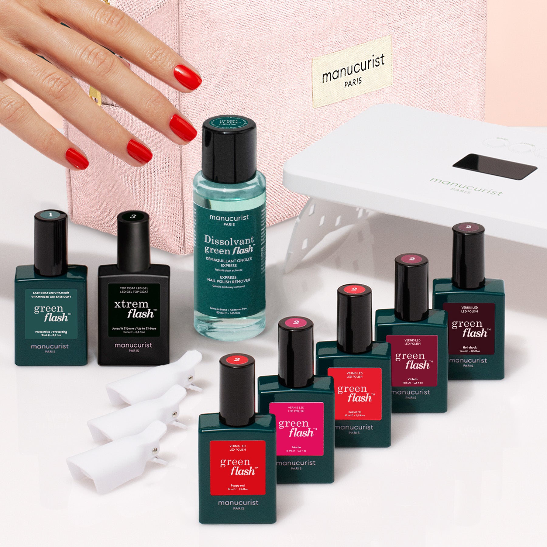 A hand with red nails reaches towards the Manucurist Super Starter Kit—featuring Green Flash gel polish, remover, a pink bag, a white LED lamp, and nail clips on a white surface—perfect essentials for achieving an ultra-glossy manicure with a vitaminized base coat.