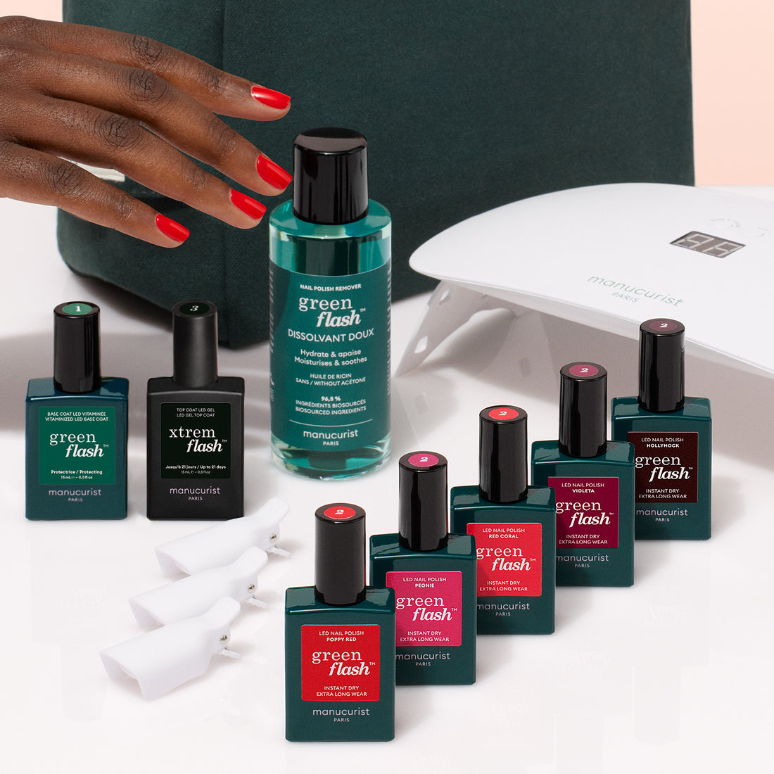 A hand with red nails reaches for the Manucurist Super Starter Kit, featuring Green Flash manicure essentials like a Vitaminized Base Coat and nail polishes, all arranged on a white surface for an ultra-glossy finish.