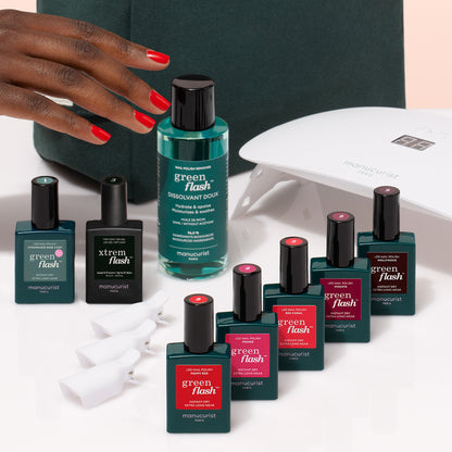 A hand with red nails reaches for the Manucurist Super Starter Kit, featuring Green Flash manicure essentials like a Vitaminized Base Coat and nail polishes, all arranged on a white surface for an ultra-glossy finish.