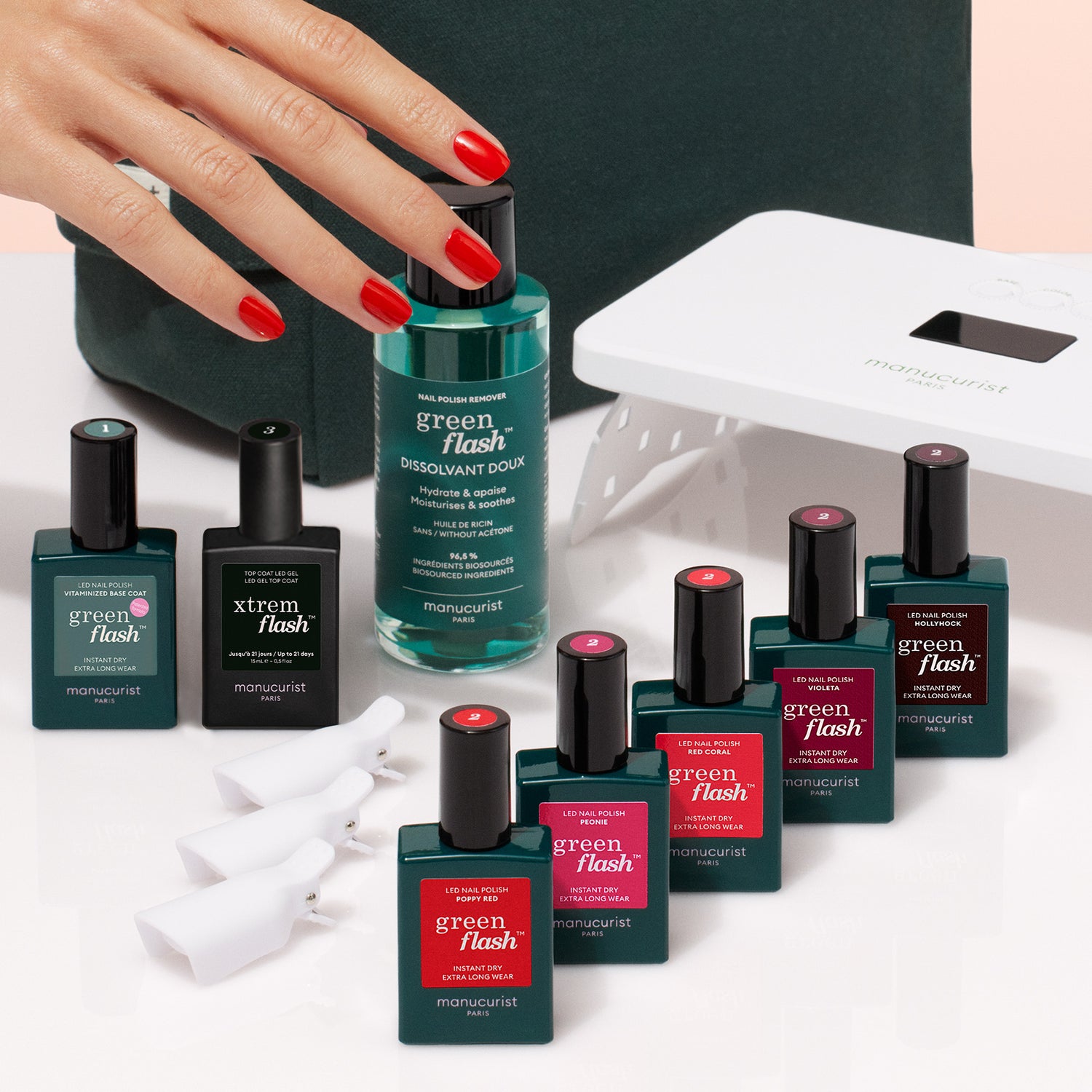 A hand with red nails reaches toward the Manucurist Super Starter Kit, featuring polishes in green and red, nail polish remover, white nail clips, a Vitaminized Base Coat, and a nail lamp on a beige surface.