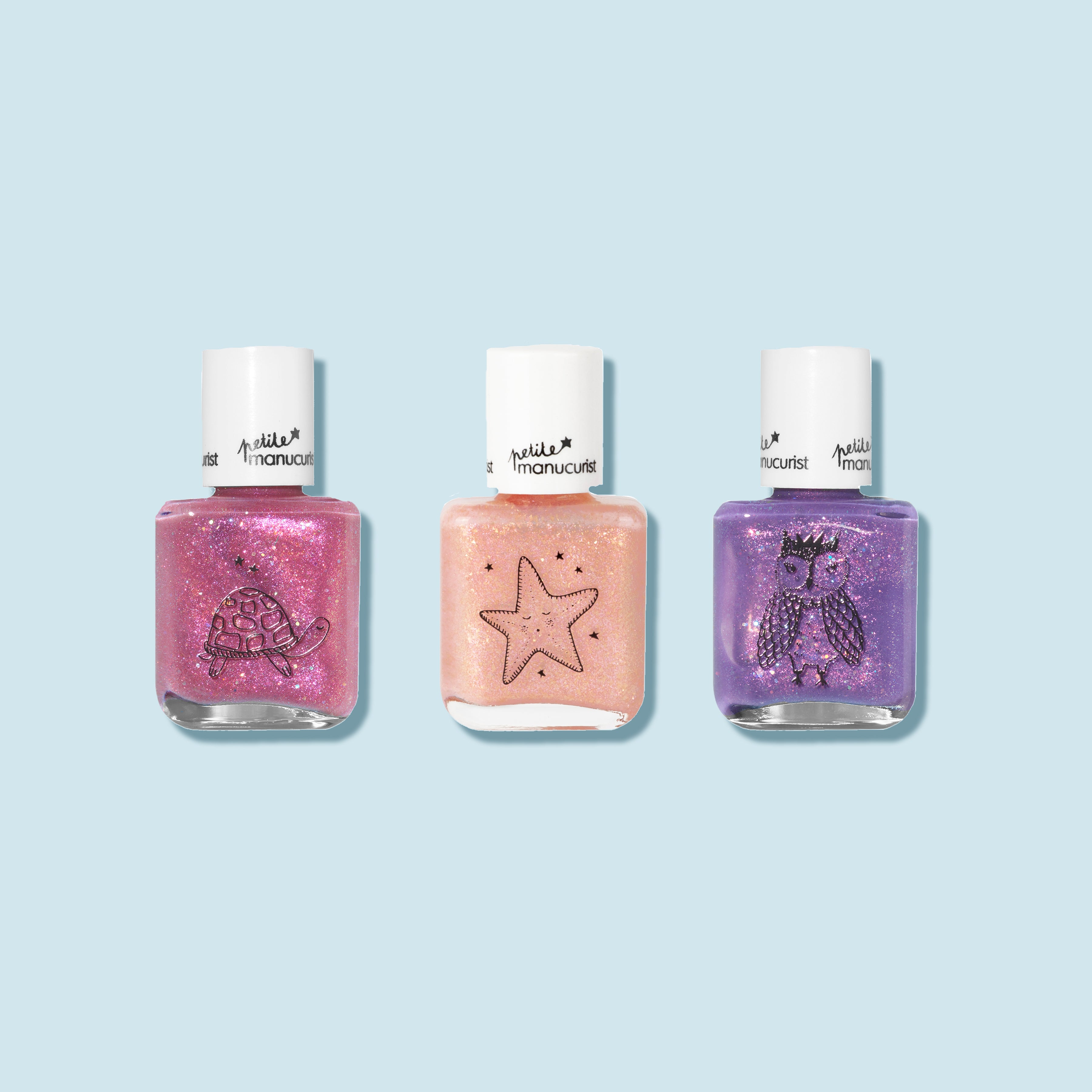 Three bottles of Manucurist’s Customisable Trio Petite Manucurist kids nail polishes with white caps on a light blue background—pink with a turtle, peach with a starfish, and purple with an owl illustration.