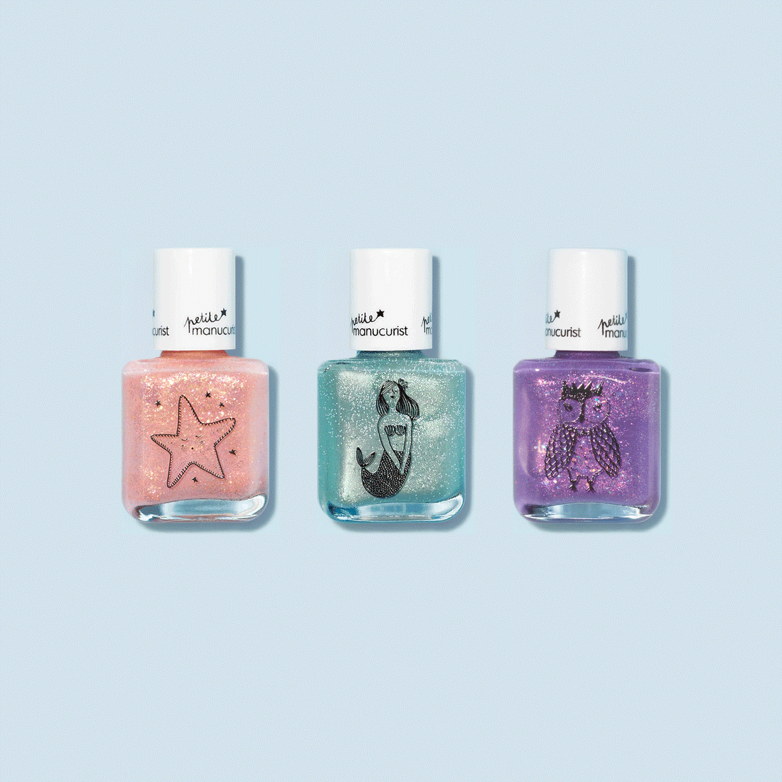 Three Customisable Trio Petite Manucurist kids nail polishes from Manucurist, each with a white cap, shine on a light blue background. The rainbow set includes pink (starfish), teal (mermaid), and purple (owl) designs.