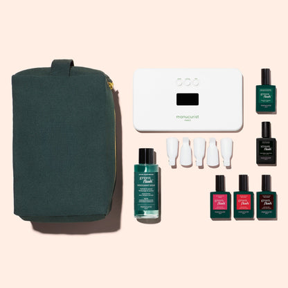 The Manucurist Starter Kit includes a dark green zippered pouch, white manicure lamp, remover, four white nail caps, and six bottles—one is a vitaminized base coat—for an ultra-glossy manicure on a pale pink background.