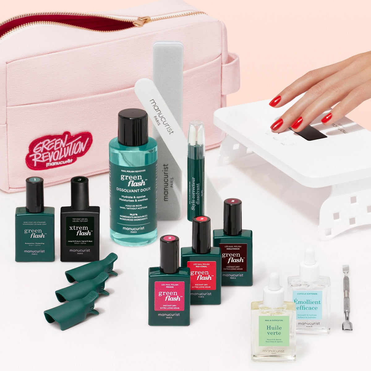 A flat lay of the Manucurist Addict Kit, featuring gel polish, a Vitaminized Base Coat, remover, nail files and tools, green finger clips, a small pink bag, and a UV lamp, with a hand receiving an ultra-glossy red gel manicure.