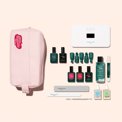 The Manucurist Addict Kit, featuring a pink zippered pouch filled with essential manicure tools—including nail polishes, files, a cuticle pusher, Vitaminized Base Coat, gel remover, and a white LED nail lamp—is neatly arranged on a pale background, offering everything you need for an ultra-glossy manicure.