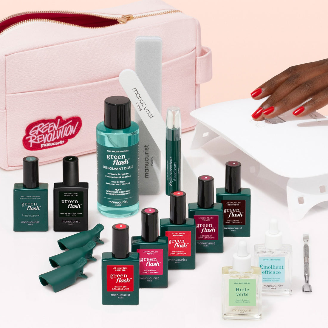 A hand with red nails is near a UV lamp. The Manucurist Super Addict Kit, featuring various green and pink manicure bottles—including Green Flash gel polish and Vitaminized Base Coat—along with nail tools and a pink bag labeled “Green Revolution,” is arranged on a white surface for an ultra-glossy manicure.