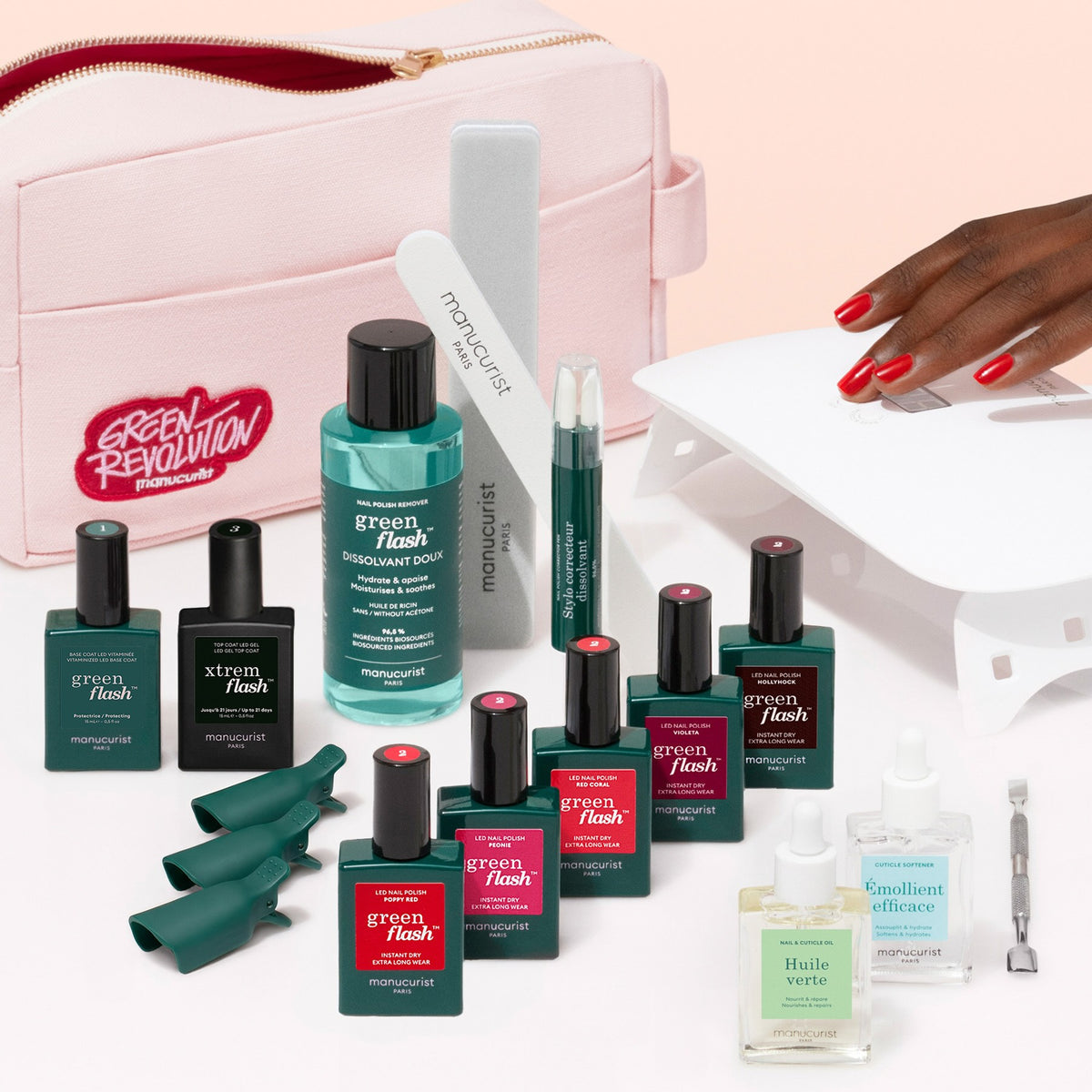 A hand with red nails is near a UV lamp. The Manucurist Super Addict Kit, featuring various green and pink manicure bottles—including Green Flash gel polish and Vitaminized Base Coat—along with nail tools and a pink bag labeled “Green Revolution,” is arranged on a white surface for an ultra-glossy manicure.