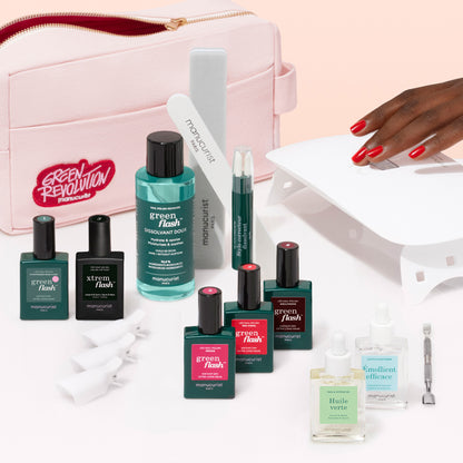 A pink pouch labeled Manucurist Addict Kit sits among nail care essentials—Green Flash gel polish, remover, vitamin base coat, nail files, cuticle oil, a nail lamp—and a hand with ultra-glossy red nails touching the lamp.