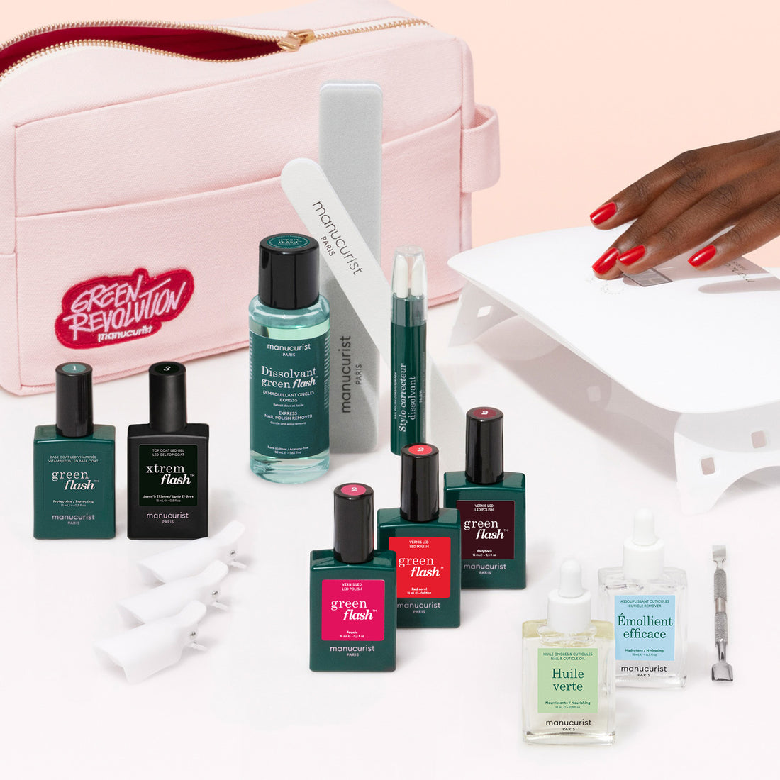 A hand with red polished nails, prepped with Manucurist Addict Kit, reaches under a white lamp. Around it are gel polish bottles, manicure tools, nail care items, and a pink bag featuring the Green Revolution slogan.