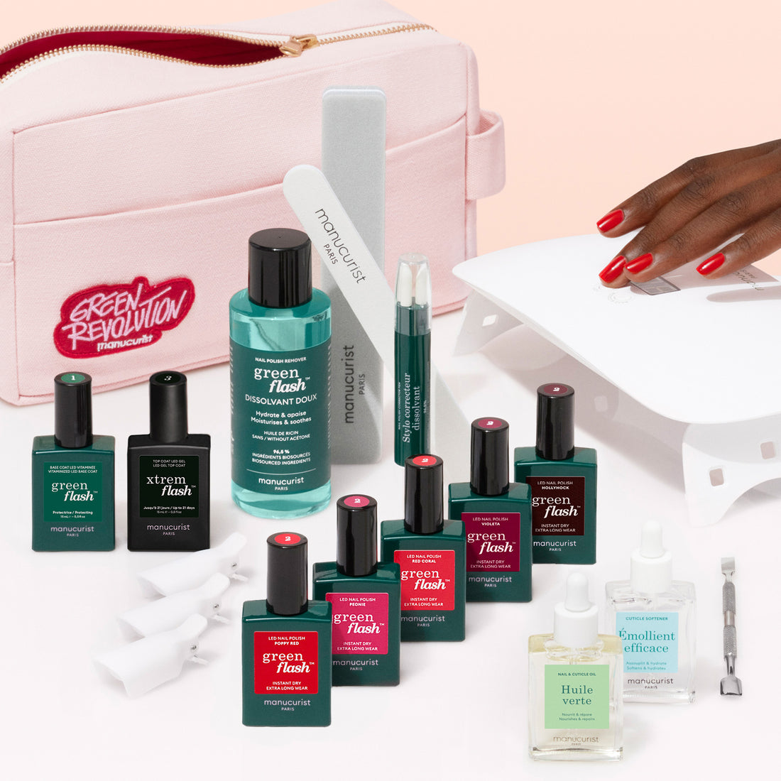 A hand with red nails rests on a white UV lamp, surrounded by Manucurist Super Addict Kit products—gel polish bottles, vitamin-enriched base coat, nail care tools, oils, and a pale pink Green Revolution case—all neatly arranged.