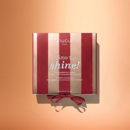 A red and gold box labeled “Manucurist Advent Calendar Time to Shine!” featuring a red ribbon, ideal for nail care enthusiasts, is displayed on a gradient peach background.