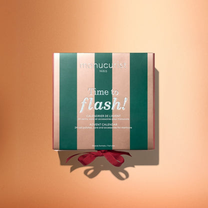 A box featuring pink and green stripes, labeled “Manucurist Advent Calendar Time to Flash!”, ideal for gel polish manicure enthusiasts, rests on a peach background with a red ribbon at the bottom.