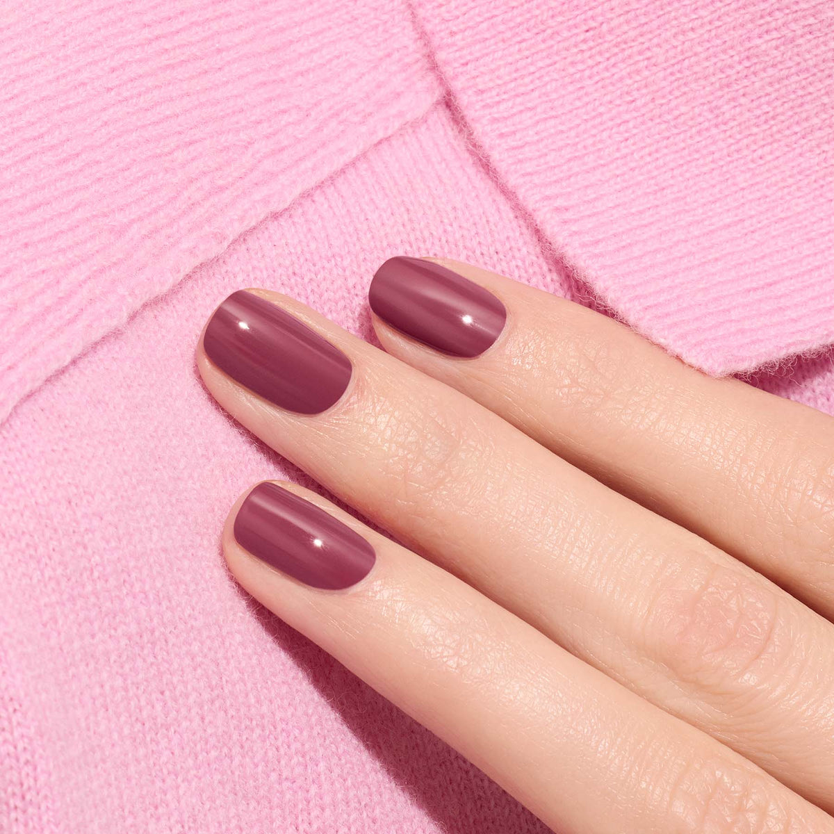 A close-up of neatly manicured nails painted in Manucurist’s Victoria Plum, a glossy plum shade, resting on light pink textured fabric.
