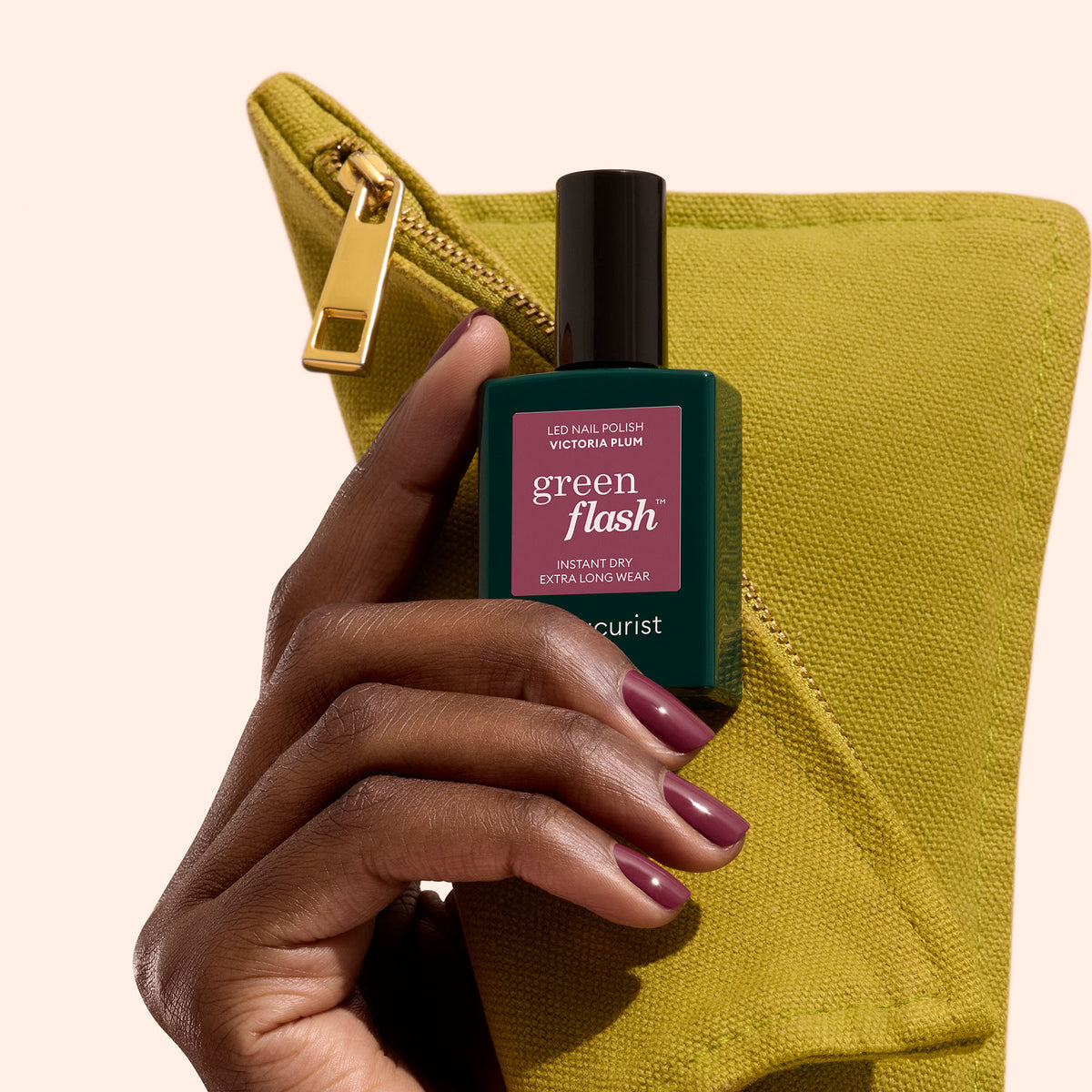 A hand with Manucurist Victoria Plum nail polish holds a green Green Flash gel polish bottle in front of a yellow-green zip pouch, set against a light beige background.