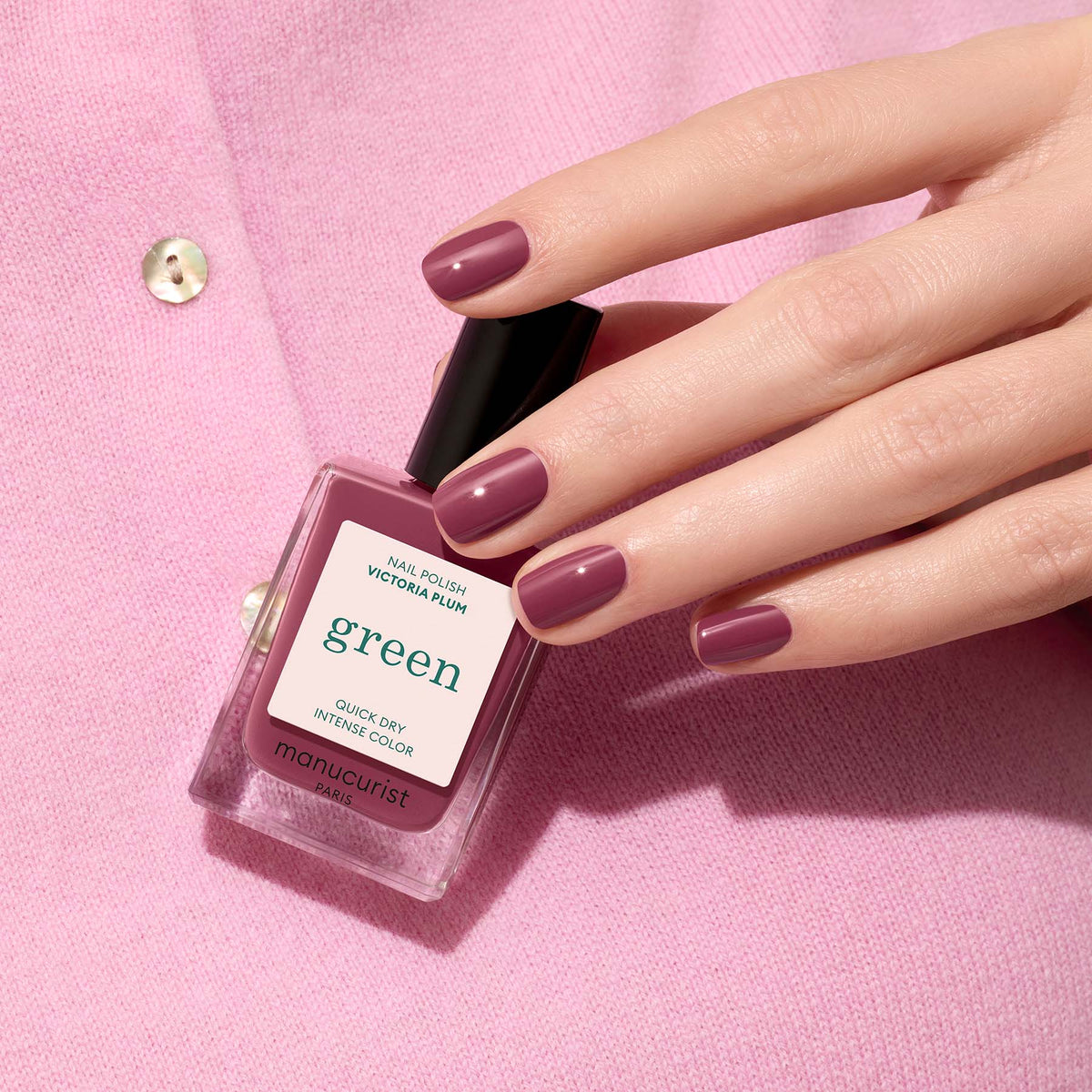 A hand with Victoria Plum nail polish holds a matching Manucurist bottle against a pink sweater with gold buttons.