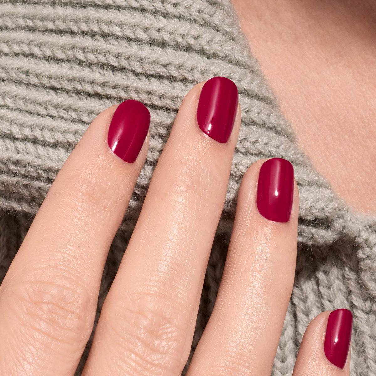 A close-up of a hand with neatly manicured nails painted in Manucurist’s Violeta, a glossy burgundy purple shade, resting on light gray knitted fabric.