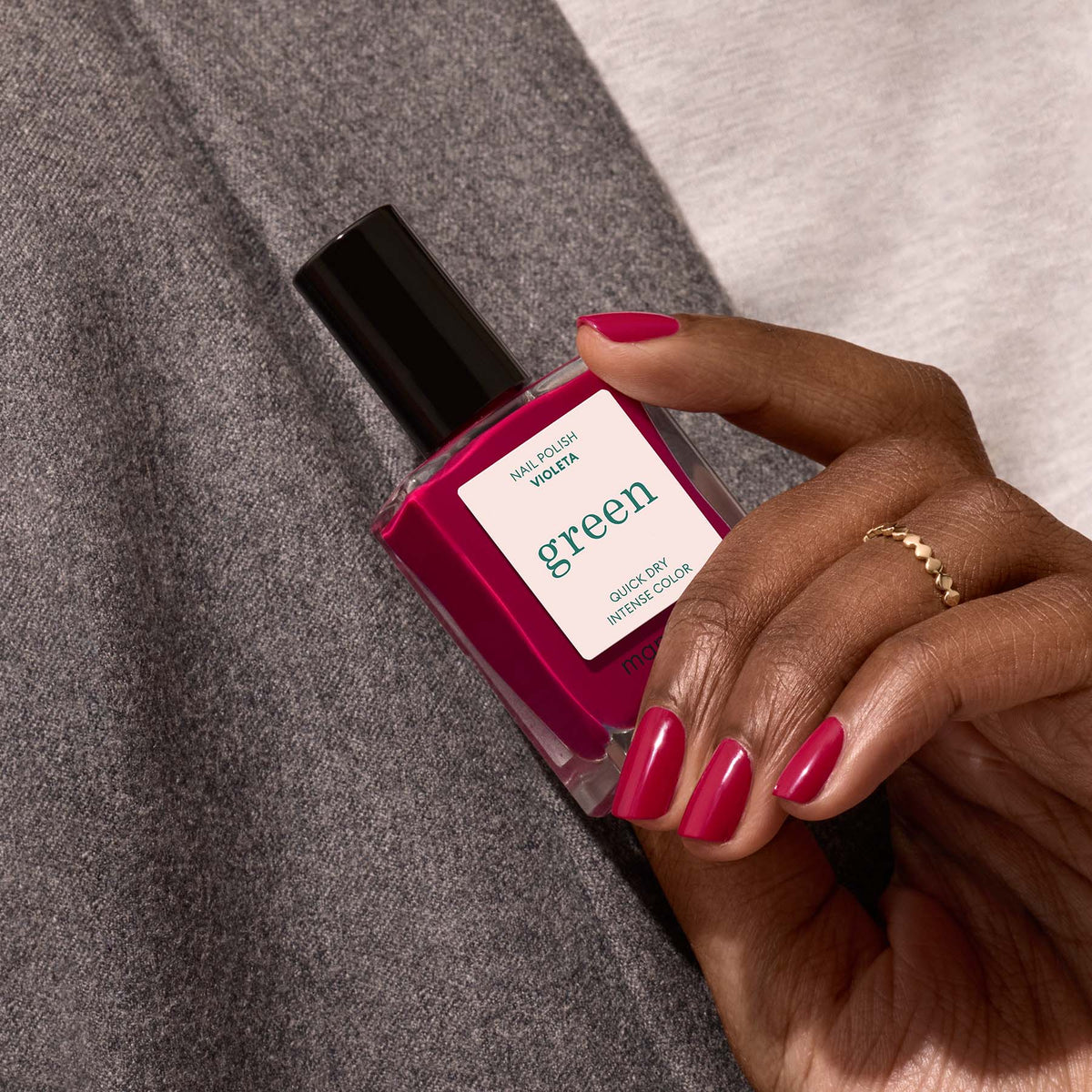 A hand with neatly manicured pink nails holds a bottle of Manucurist Violeta nail polish against a grey and white fabric background. The person wears a gold ring on their index finger.