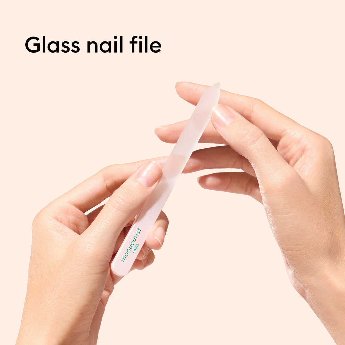 A person shapes their fingernail with a Manucurist Glass nail accessory trio. The product name appears at the top left. A glass cuticle pusher is nearby, set against a soft beige background.