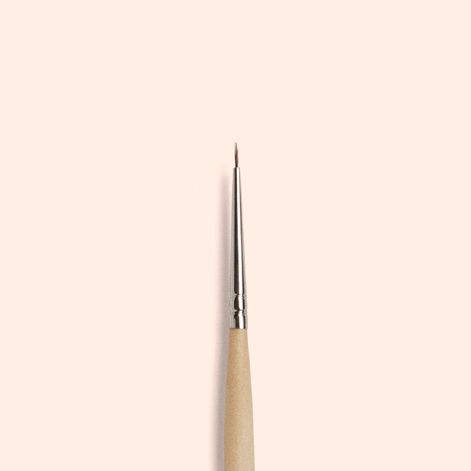 The Manucurist Fine Tip Brush, made with vegan synthetic bristles and a wooden handle, stands vertically against a light beige background—ideal for precision nail art.