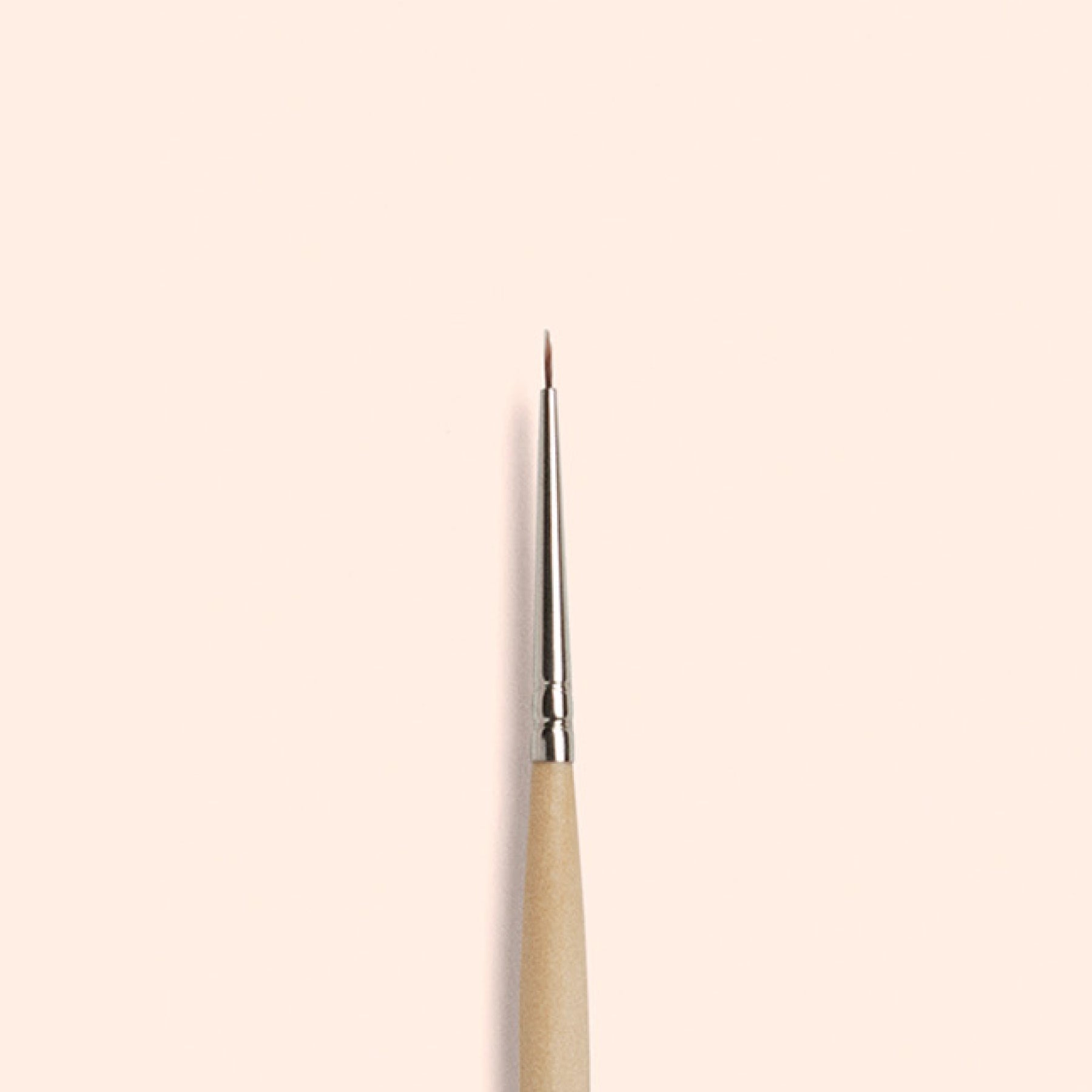 The Manucurist Fine Tip Brush, made with vegan synthetic bristles and a wooden handle, stands vertically against a light beige background—ideal for precision nail art.