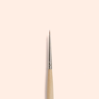 The Manucurist Fine Tip Brush, made with vegan synthetic bristles and a wooden handle, stands vertically against a light beige background—ideal for precision nail art.