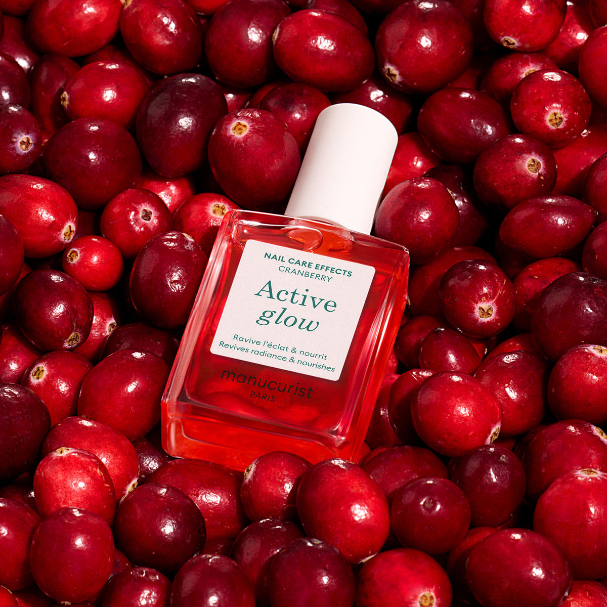 A bottle of Manucurist Active™ Glow Cranberry nail care, with a white cap and pink-toned liquid, featuring a plant-based formula, is placed on a bed of fresh cranberries.