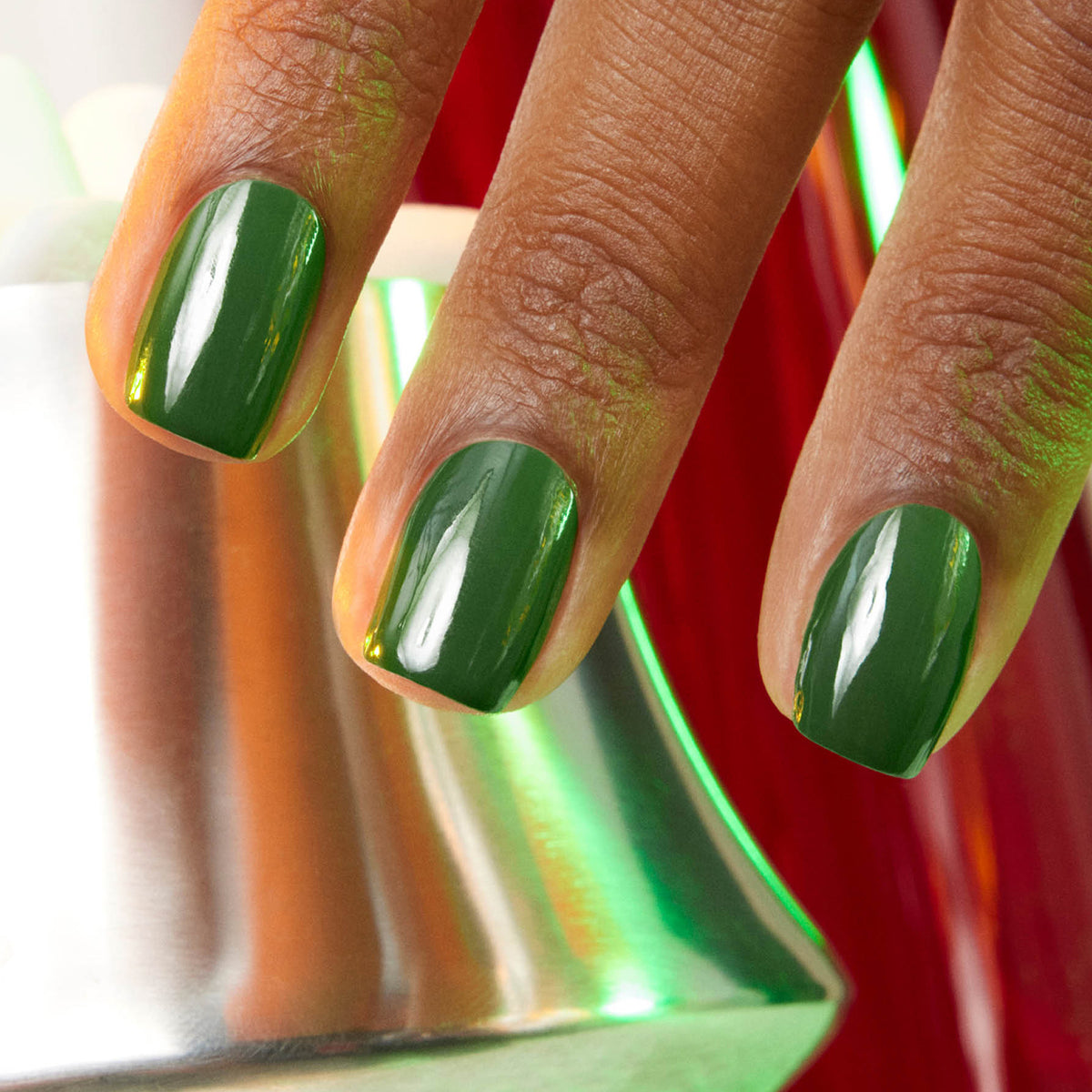 Close-up of three medium brown fingers with Manucurist Jade, a glossy mineral green polish, held near a metallic object reflecting green and red light. The cool undertones of the vibrant shade complement the skin tone.