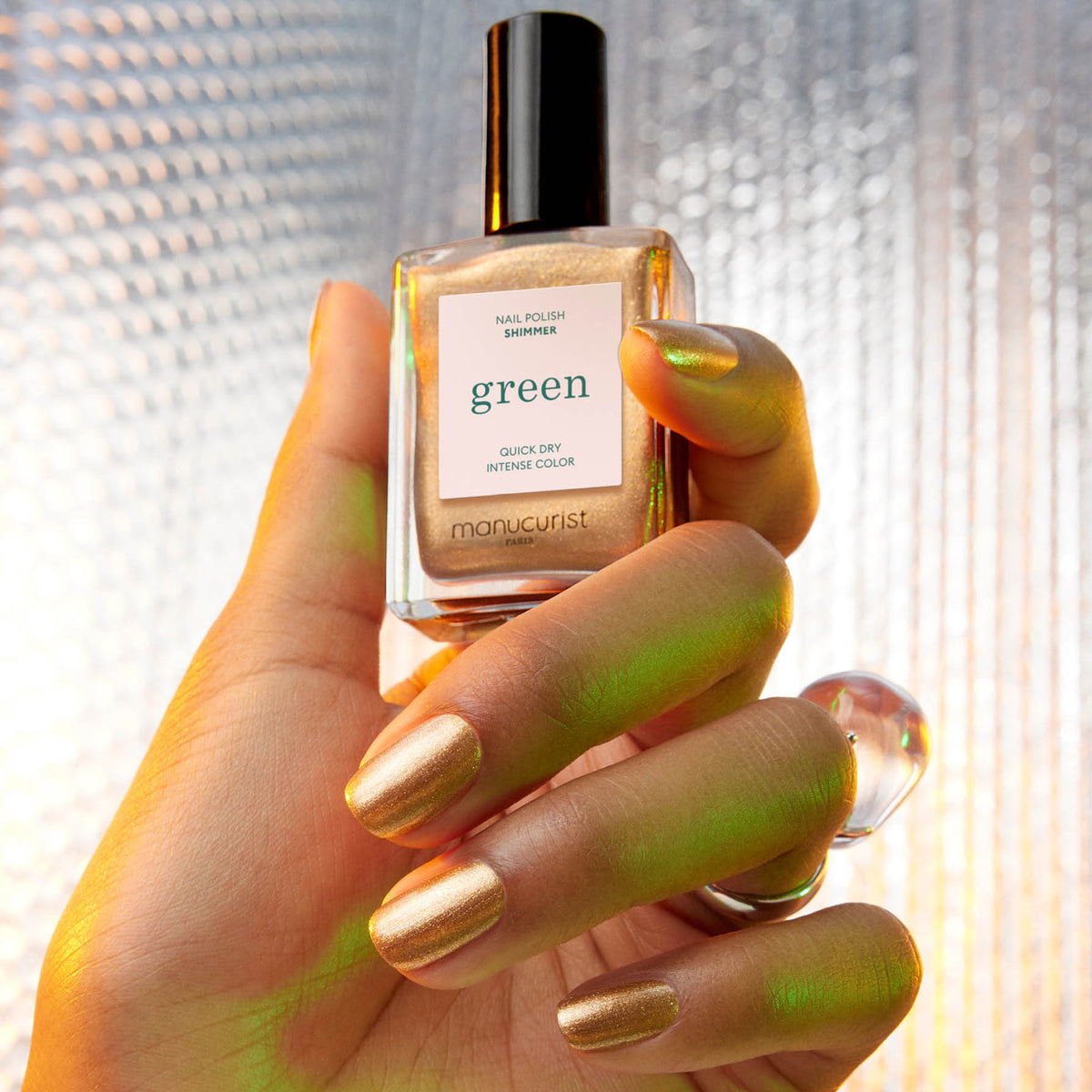 A hand with metallic gold nail polish featuring cool undertones holds a matching bottle labeled "Shimmer" by Manucurist against a textured, light-reflective background.