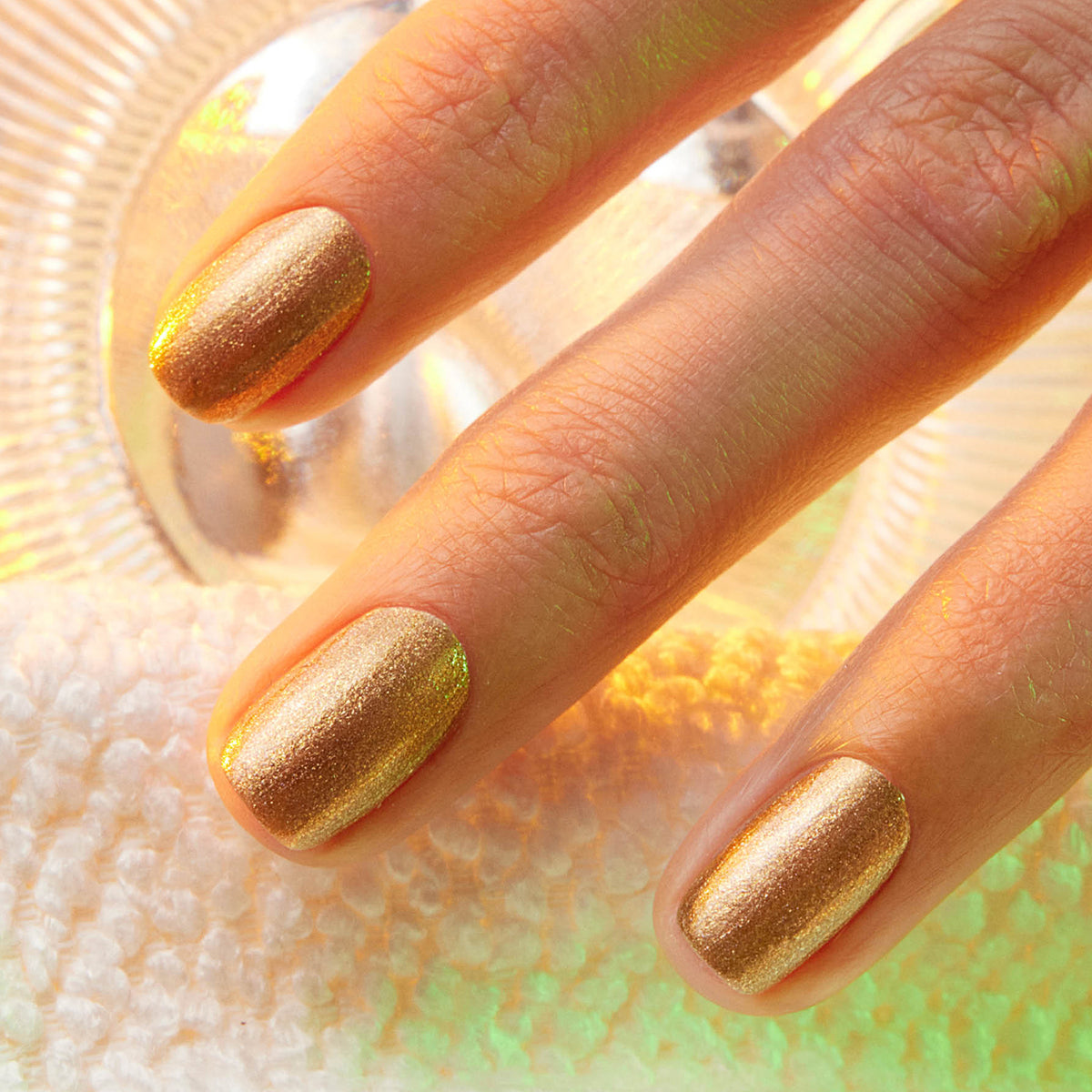 A close-up of three fingers with short, neatly manicured nails painted in Manucurist’s Shimmer polish. The hand rests on a textured white surface, bathed in warm, glowing light in the background.