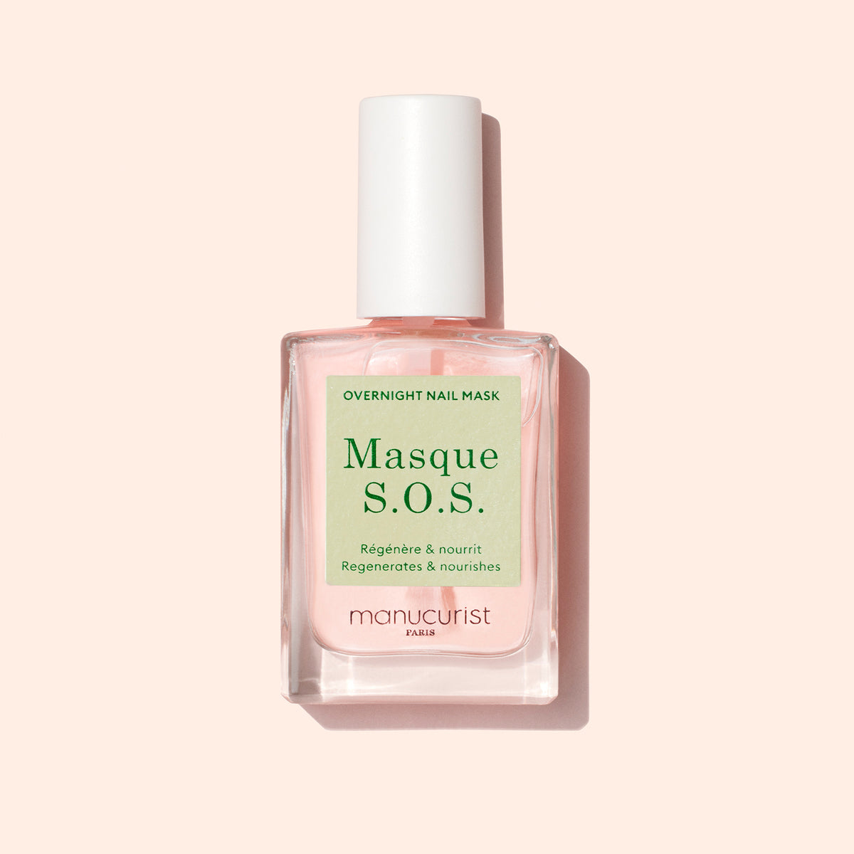 A bottle of Manucurist S.O.S. Nail Mask with a white cap and pale pink liquid sits on a light pink background. The label has green and white text. This overnight mask is perfect for nail regeneration.
