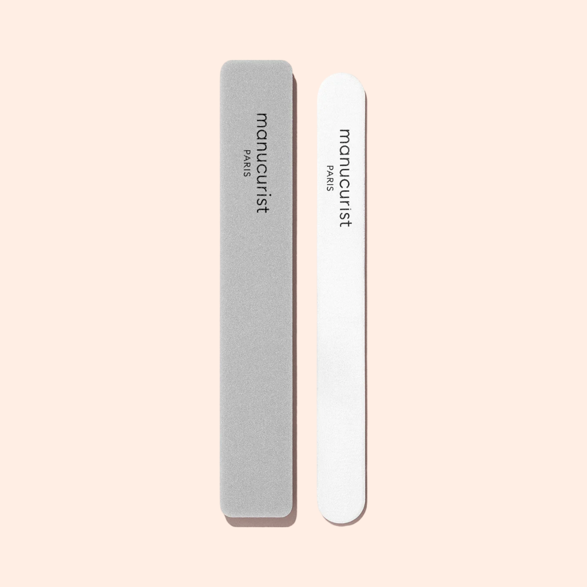 Two Manucurist Nail buffer sponges are placed side by side on a light beige background—one grey and rectangular, the other white and rounded. Both feature “manucurist PARIS” printed on them.