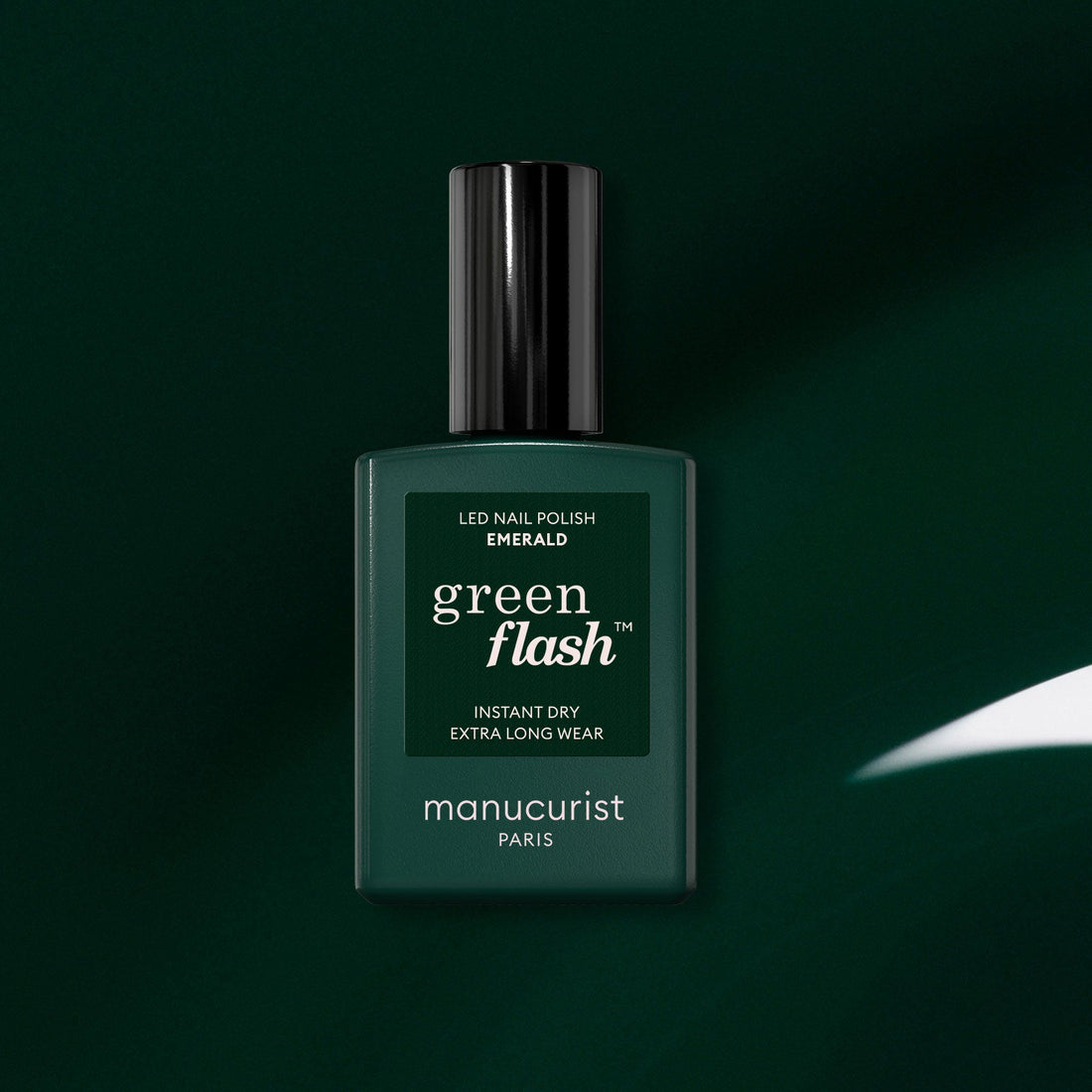 A dark green bottle of Manucurist Green Flash LED gel nail polish in the shade Emerald is displayed against a matching green background, with a white reflection highlighting its deep, gemstone-inspired color.