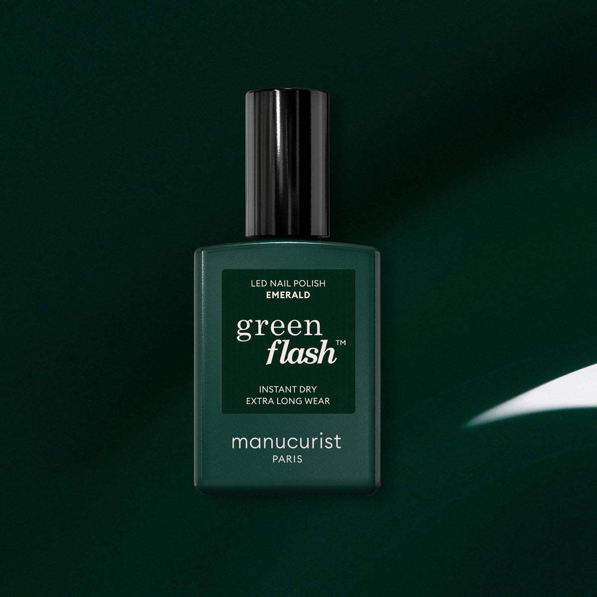 A dark green bottle of Manucurist Green Flash LED gel nail polish in the shade Emerald is displayed against a matching green background, with a white reflection highlighting its deep, gemstone-inspired color.