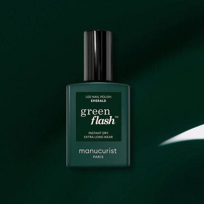 A dark green bottle of Manucurist Green Flash LED gel nail polish in the shade Emerald is displayed against a matching green background, with a white reflection highlighting its deep, gemstone-inspired color.