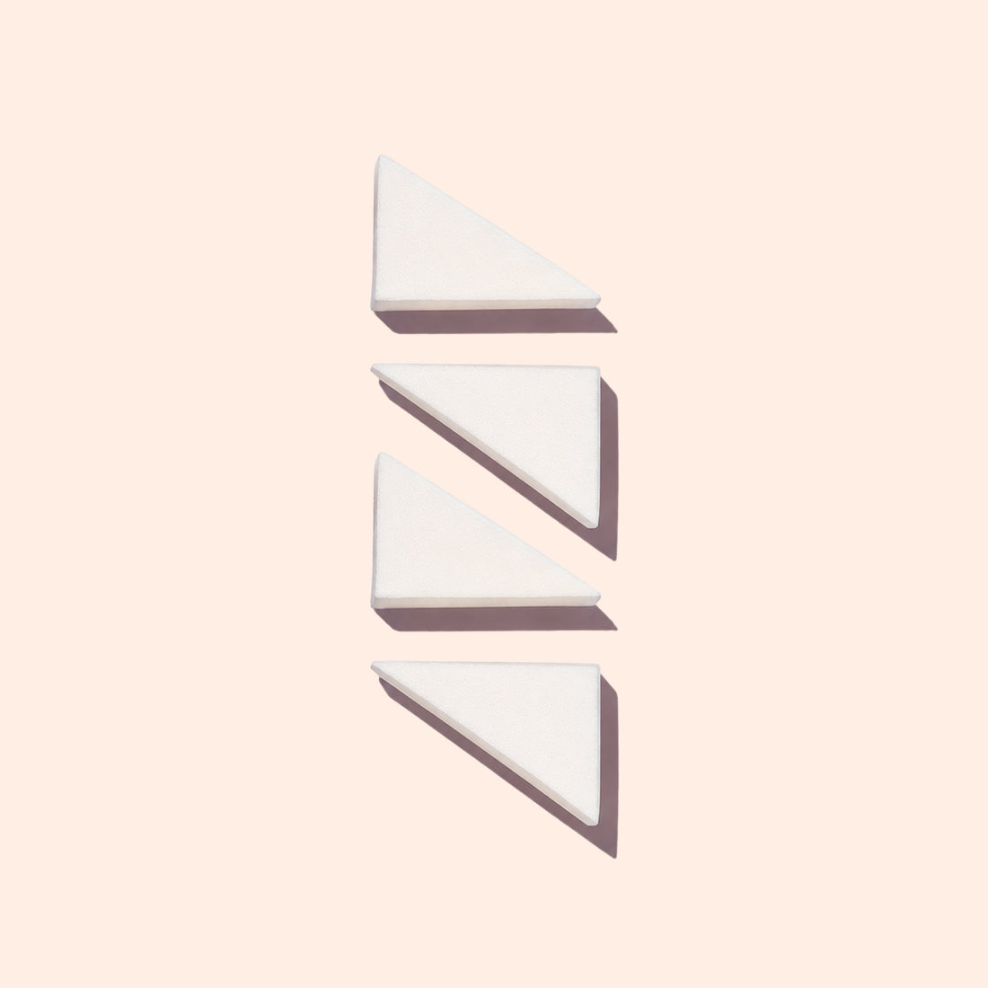 Three white triangular wedges from Manucurist’s Nail Art Sponges - x4 are arranged vertically on a light beige background, each casting a shadow to the lower right.