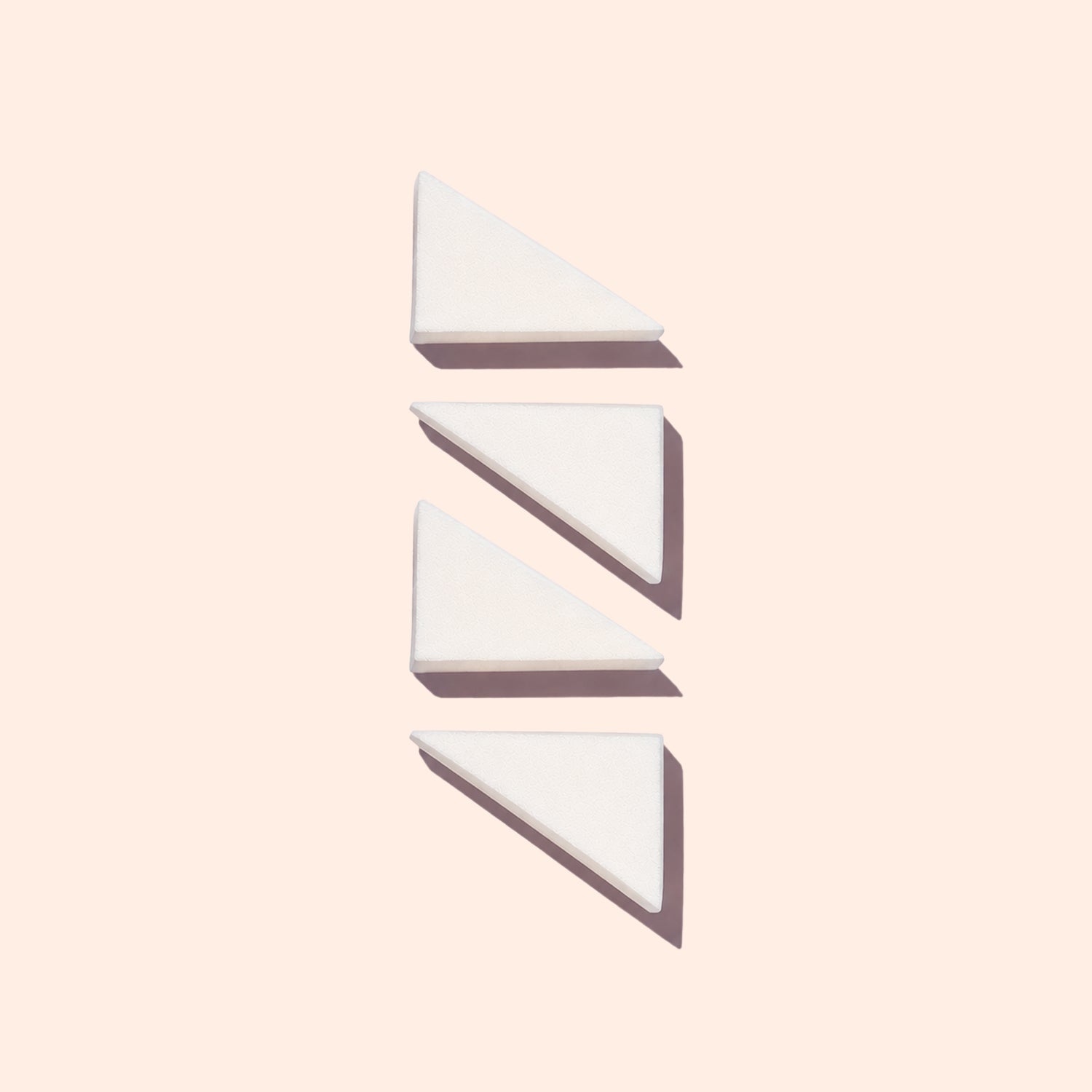 Three white triangular wedges from Manucurist’s Nail Art Sponges - x4 are arranged vertically on a light beige background, each casting a shadow to the lower right.
