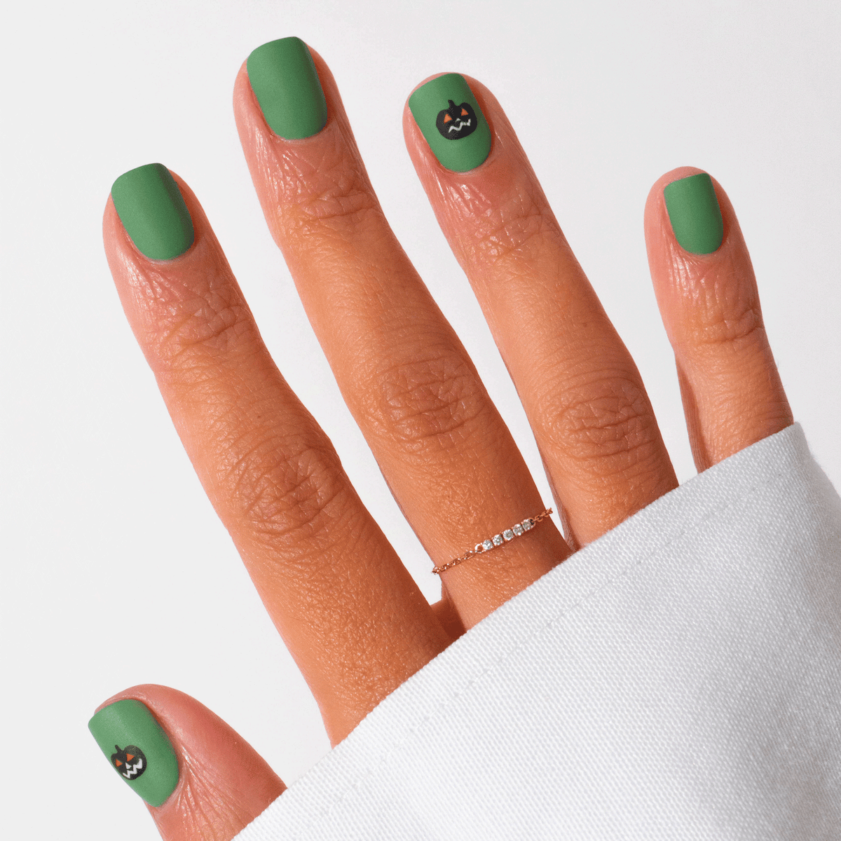 A hand with Manucurist’s Pumpkin Patch matte green gel polish displays a small black cat face on the pinky and middle nails. A delicate gold ring adorns the hand, partially covered by a white sleeve.