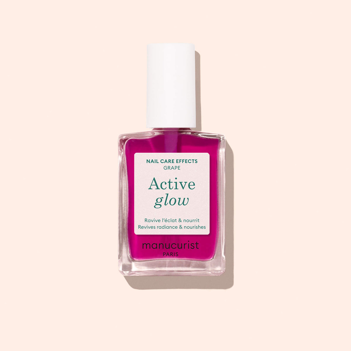 A clear glass bottle of Manucurist Active™ Glow Grape nail care polish with a white cap, filled with bright pink liquid that delivers a natural shine, is shown against a pale beige background.