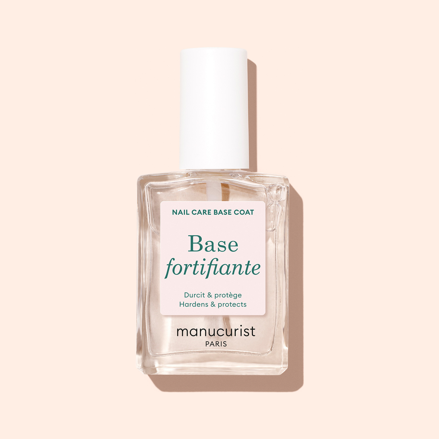A clear glass bottle of Manucurist Force Nail Hardener with a white cap is shown against a pale beige background. The label reads, "Hardens  protects.